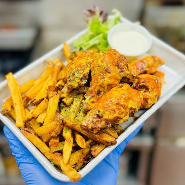 Basket of saucy chicken wings and seasoned fries with dipping sauce, held by a gloved hand in a restaurant kitchen.