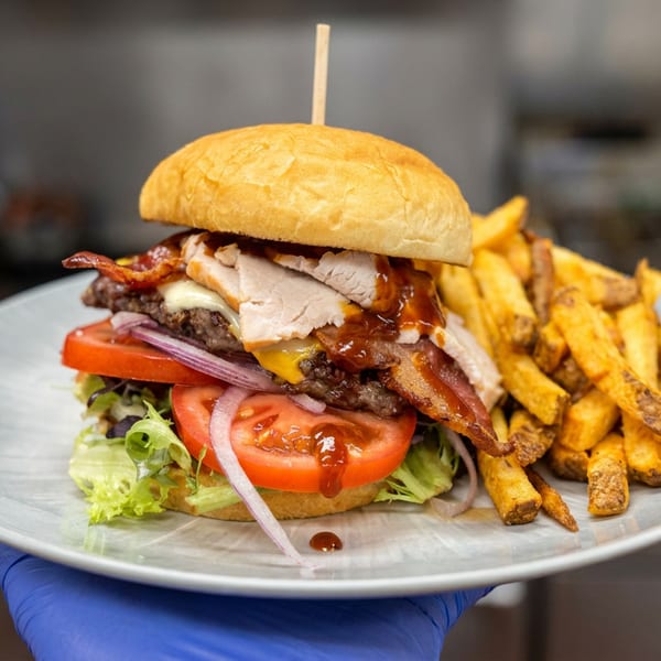 Loaded burger with bacon, turkey, cheese, tomatoes, lettuce, and onions served with golden fries on a plate.