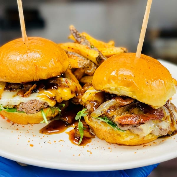 Two gourmet cheeseburgers with glossy buns, caramelized onions, greens, and sauce served on a white plate.