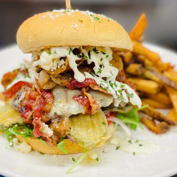 Loaded gourmet burger with melted cheese, bacon, crispy onions, sauce, and fries served on a white plate.