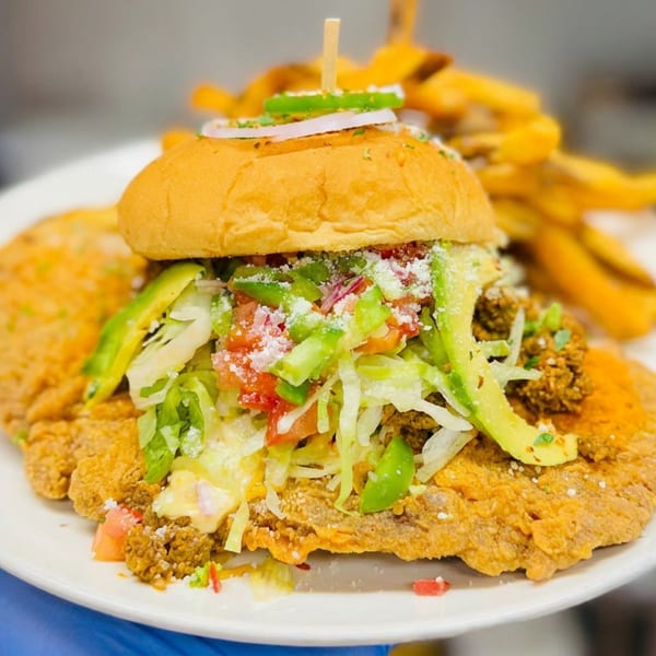 Crispy fried chicken sandwich topped with lettuce, peppers, tomatoes, onions, and cheese, served with fries on a white plate.